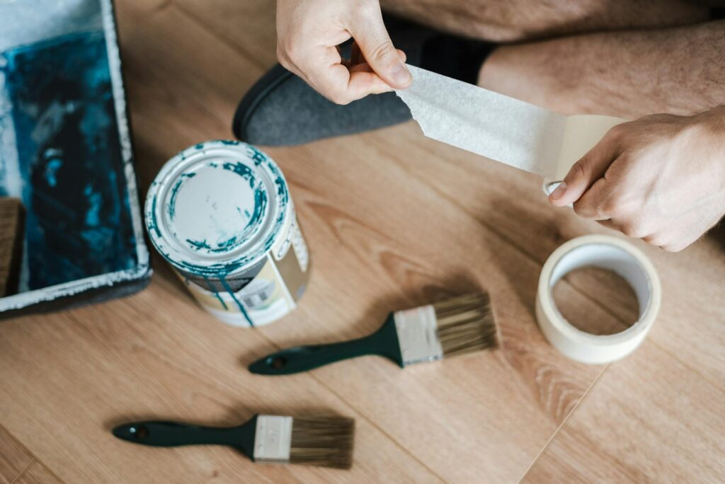 From above of crop anonymous male unwrapping sticky tape roll on parquet near paint brushes in house